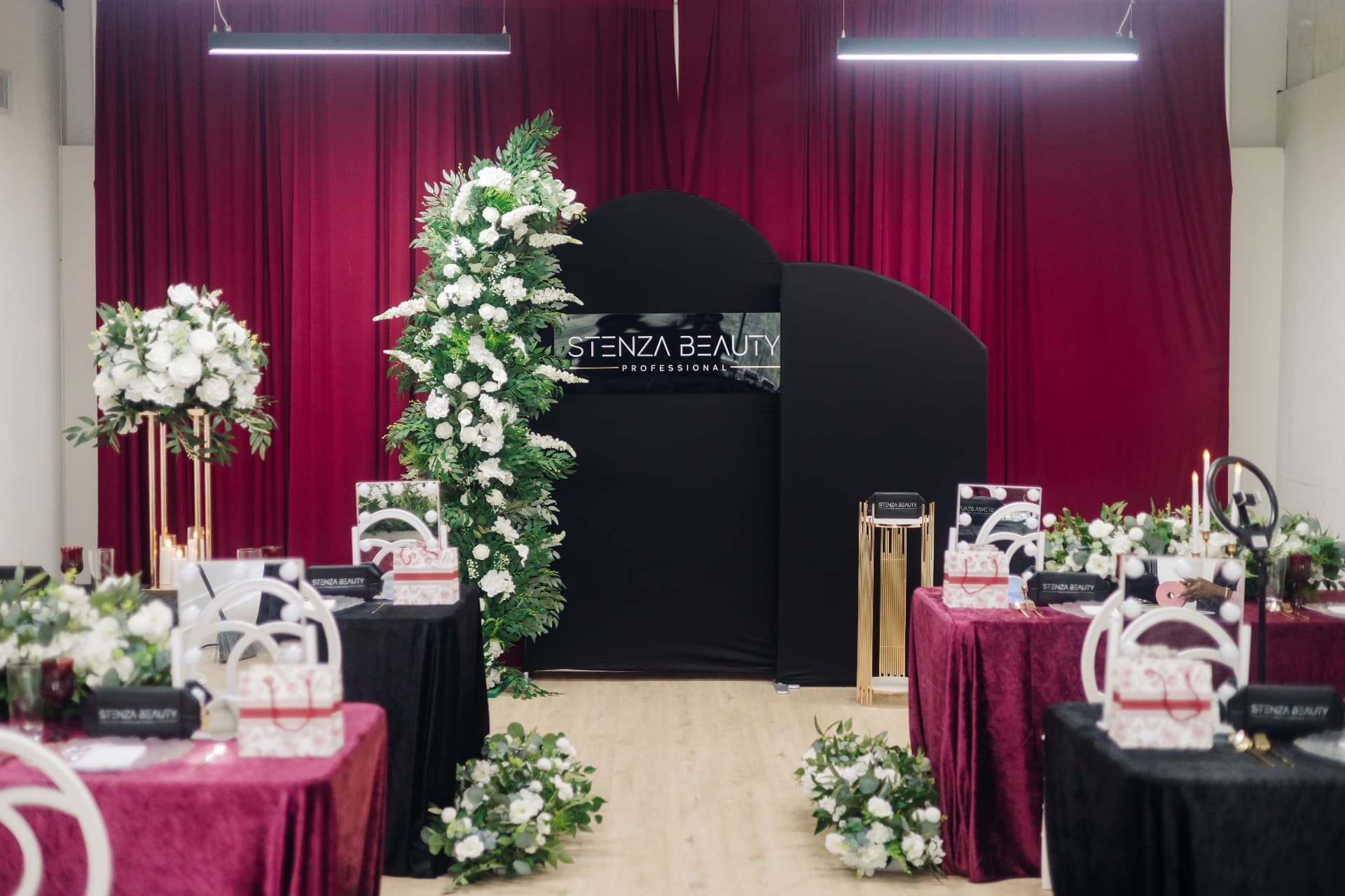 Black arched Stenza Beauty backdrop above red velvet tables and a tall floral tower