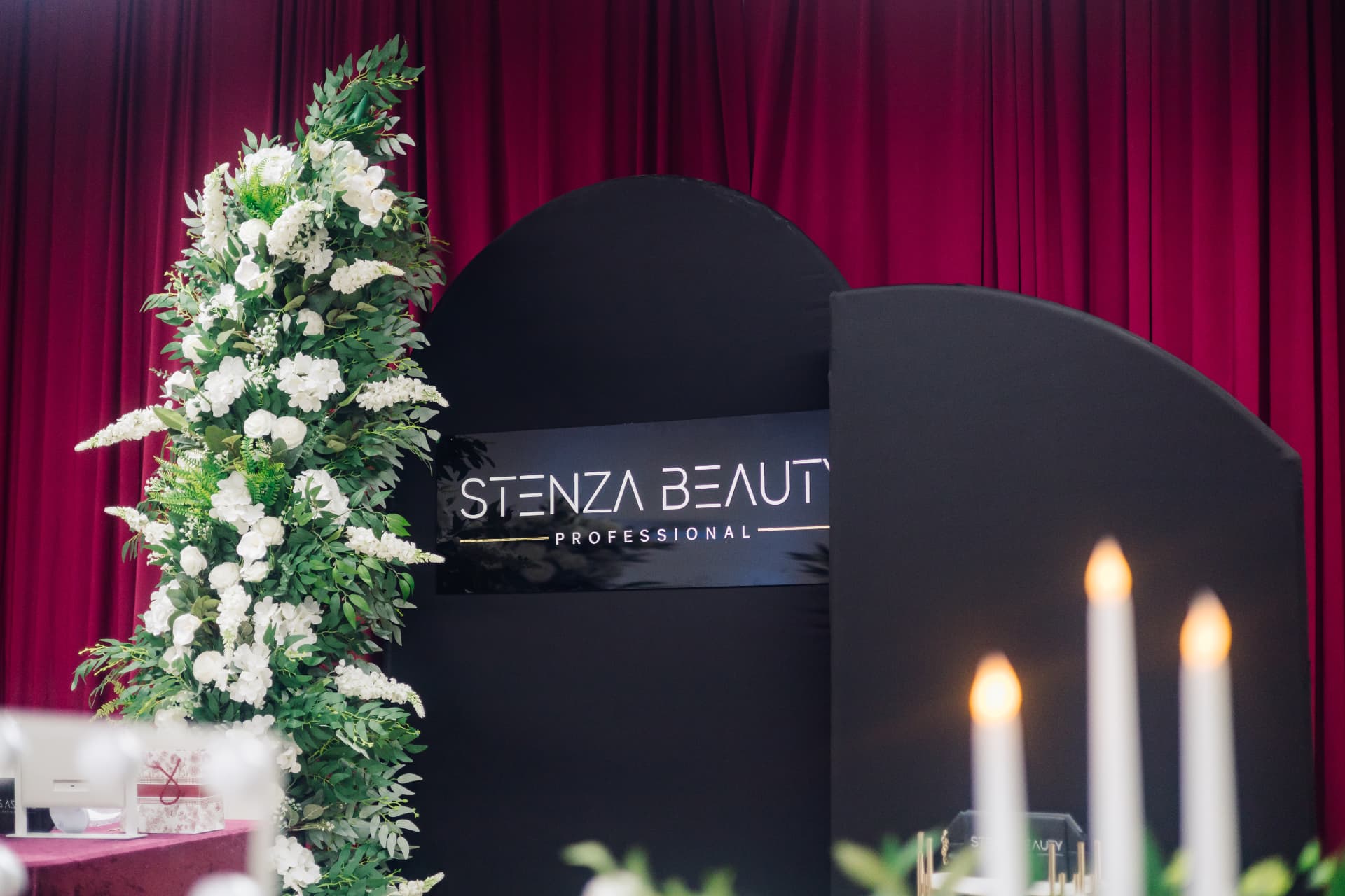 Stenza Beauty backdrop framed by white roses and greenery against red curtains